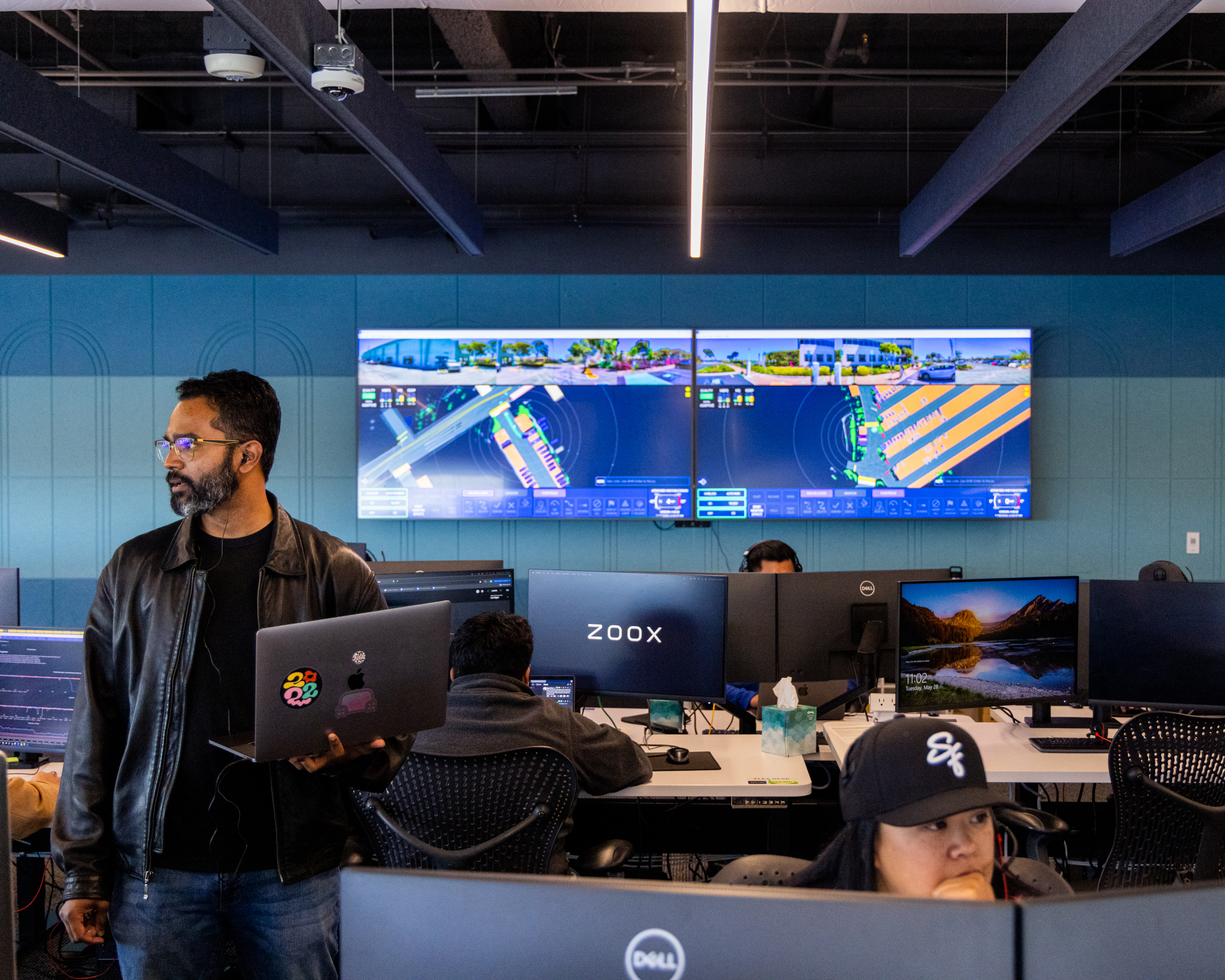 An office scene with a person standing in front of workstations with people seated at computers.