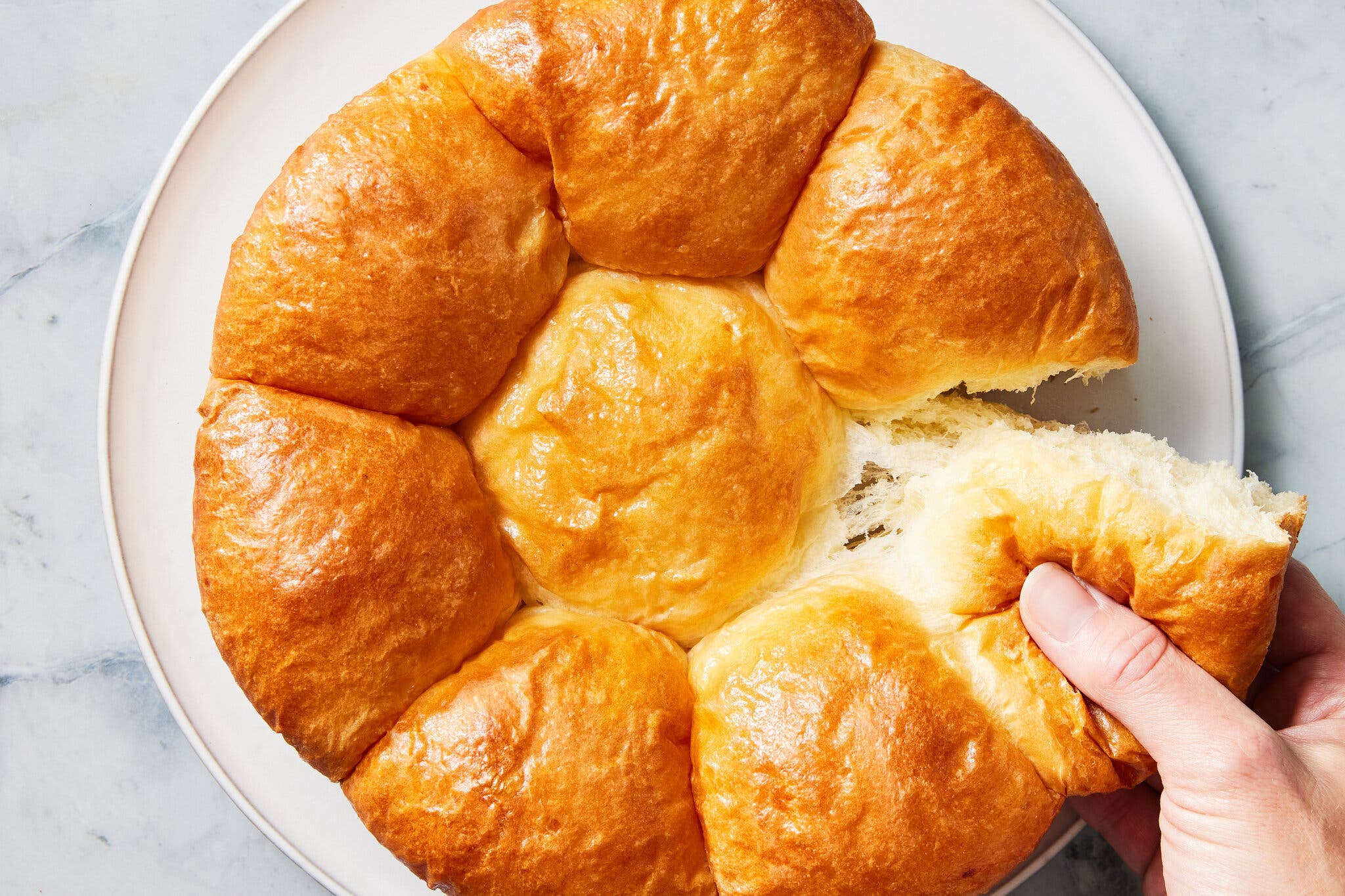 A white plate with a round loaf. A hand tears off one of the pull-apart rolls. 