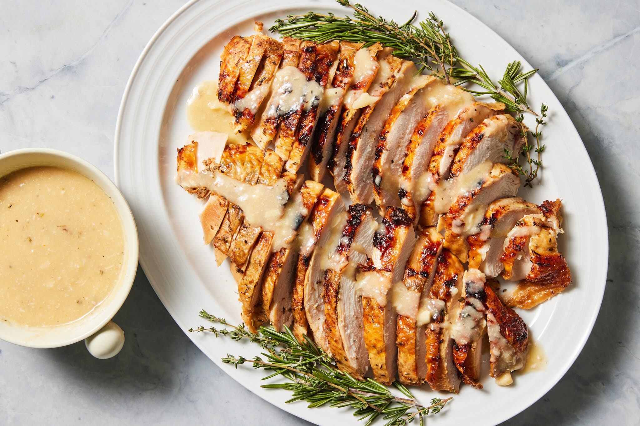 An overhead image of sliced turkey breast finished with herb sprigs and gravy on a white platter.