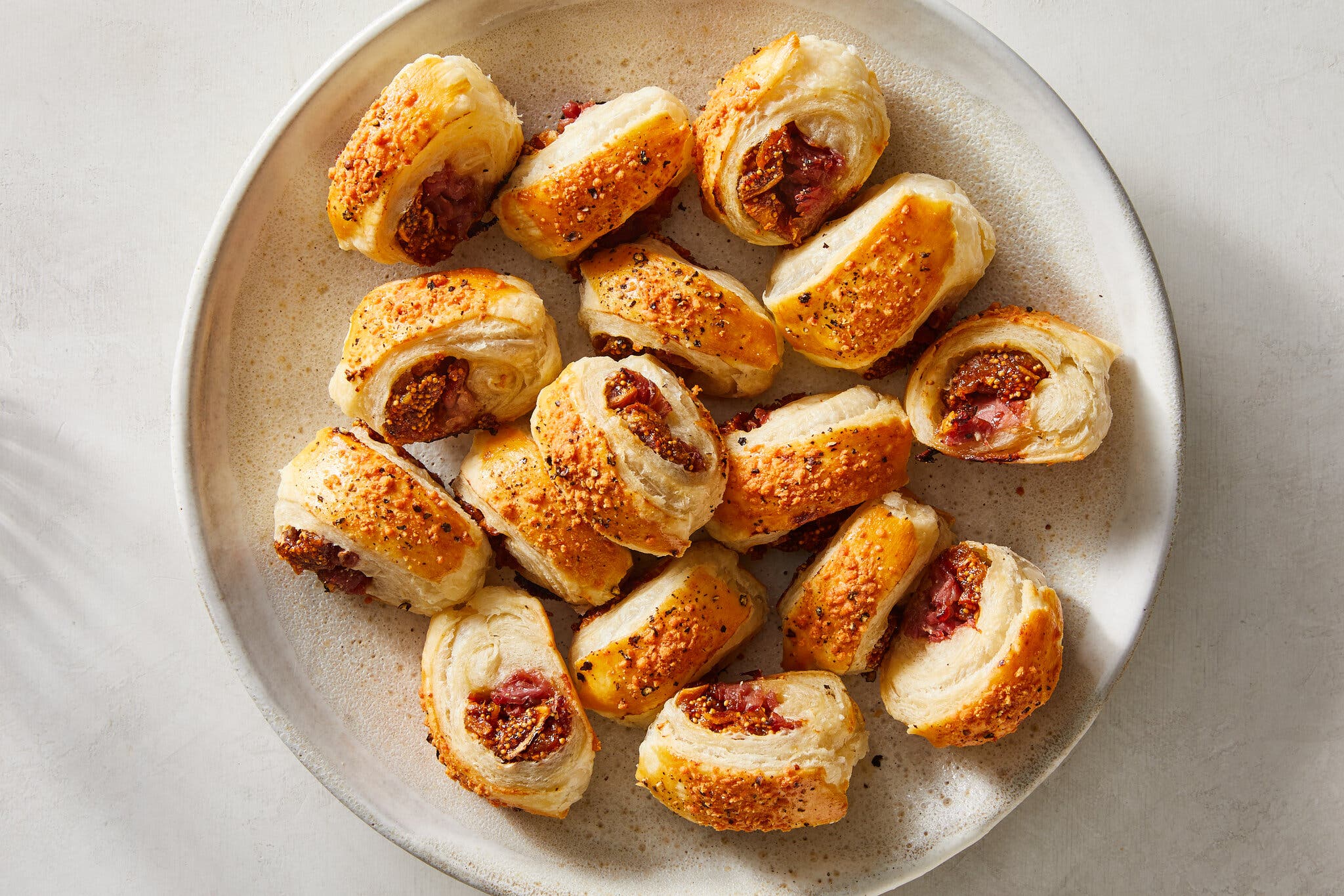 Rounds of figs and sausages wrapped with puff pastry are photographed from overhead on a white speckled platter.