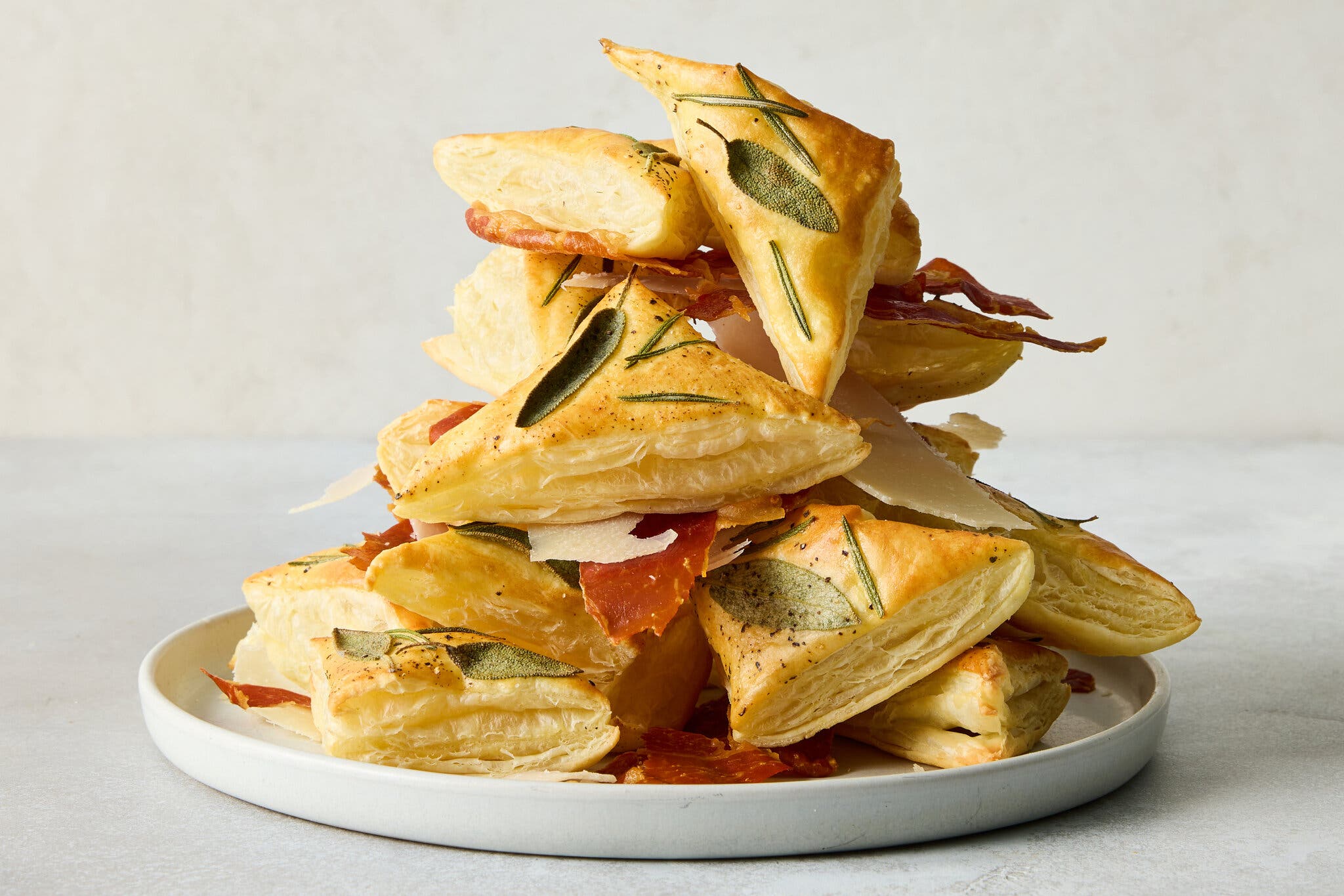 A side image of puff pastry triangles topped with herbs and sandwiching pieces of prosciutto.